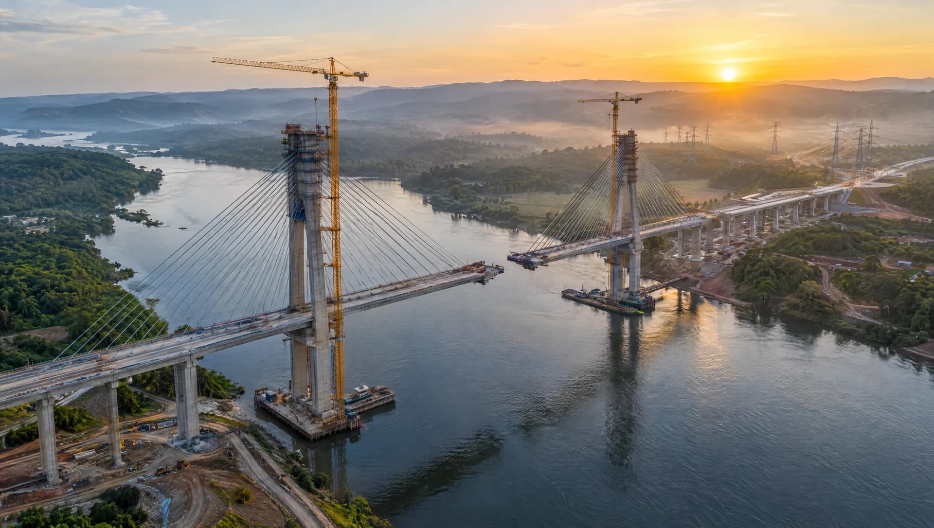 Vista aérea ao amanhecer de uma ponte estaiada em construção sobre um rio, com guindastes e linhas de transmissão