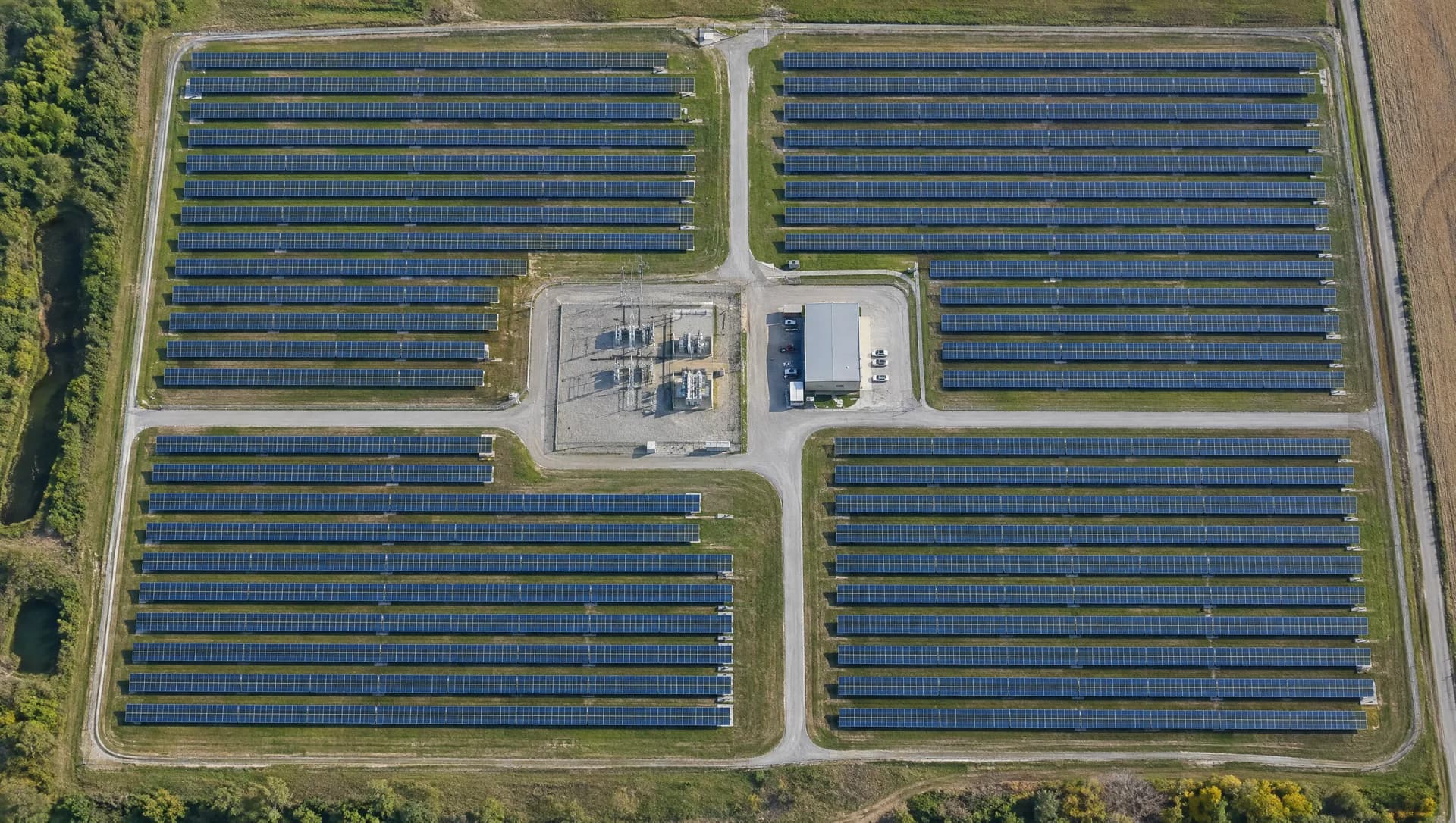 Vista aérea de cima de uma usina solar em fileiras geométricas, com vias internas, subestação e prédio de operação